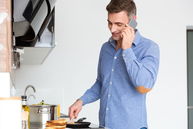 Felice bell'uomo che parla al cellulare e frigge frittelle in cucina