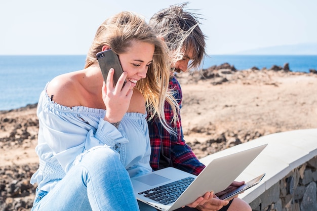 Felice bella coppia moderna che si gode la tecnologia all'aperto insieme in amicizia o relazione - giovani con laptop e telefono collegati a Internet - oceano sullo sfondo