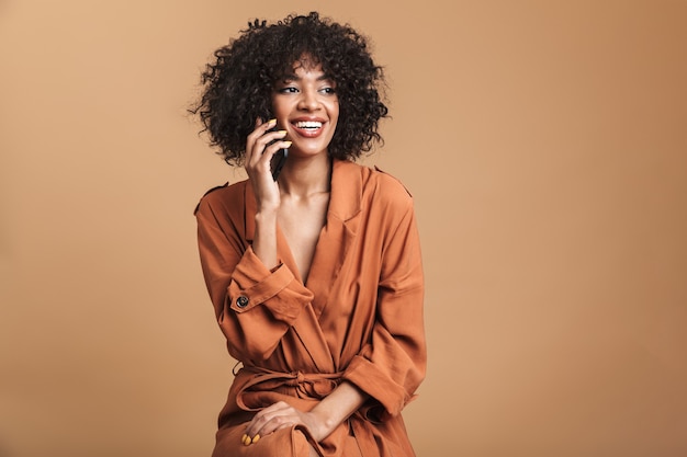 Felice bella donna africana che parla tramite smartphone e distoglie lo sguardo su sfondo marrone