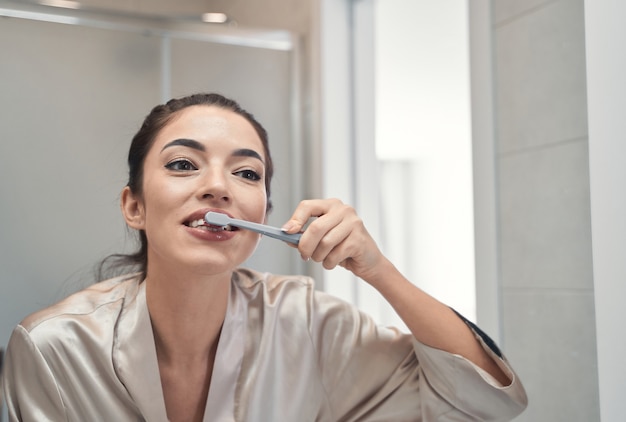 Felice bella donna con un sorriso brillante che fa le procedure mattutine in bagno mentre si lava i denti con un dentifricio