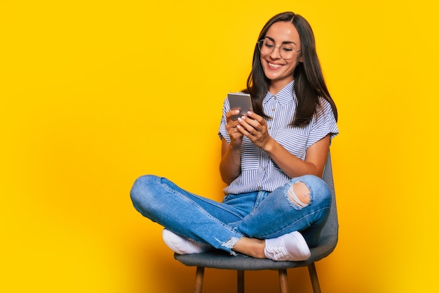 Felice bella donna elegante è seduta sulla sedia e usa il suo smartphone mentre è isolata su sfondo giallo