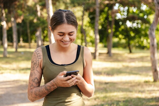 felice bella donna in abbigliamento sportivo utilizzando il cellulare e sorridente mentre si lavora nel soleggiato parco verde