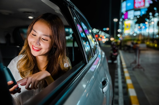 Felice bella donna sms smartphone seduto sul sedile posteriore dell'auto in città, donna d'affari asiatica che lavora fino a tardi pendolarismo dall'ufficio nel sedile posteriore del taxi con il telefono cellulare in città di notte dopo il lavoro in ritardo