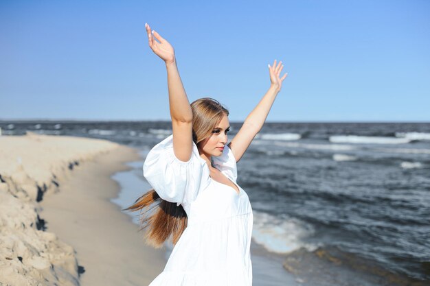 Felice, bella donna sulla spiaggia dell'oceano in piedi in un abito estivo bianco, alzando le mani.