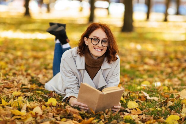 Felice bella femmina caucasica dai capelli rossi millenari in impermeabile e occhiali legge il libro giace a terra nel parco