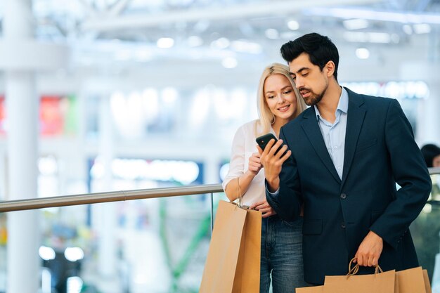 Felice bella giovane coppia utilizzando il telefono in possesso di sacchetti di carta per la spesa con gli acquisti nel centro commerciale sfondo sfocato Bell'uomo barbuto e attraente donna bionda che acquistano insieme al centro del negozio