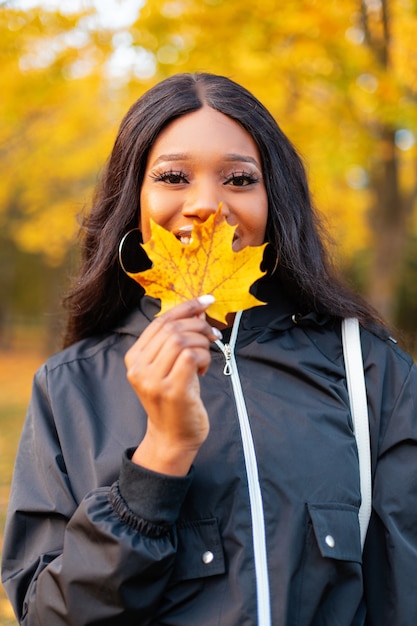 Felice bella giovane donna afroamericana con un sorriso in giacca casual nera alla moda si copre le labbra con una foglia autunnale gialla sulla natura