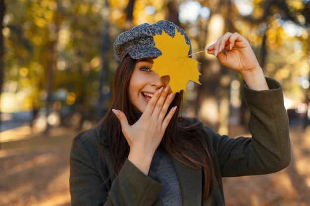 Felice bella giovane donna attraente con un sorriso in un cappotto alla moda e un cappello si copre il viso con la mano e una foglia d'autunno gialla nel parco. Emozione di gioia e meraviglia
