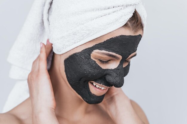 Felice bella giovane donna con maschera di argilla nera facendo terapia di benessere in posa contro la parete bianca dello studio Inquadratura orizzontale di femmina caucasica che applica maschera facciale organica Concetto di salute di bellezza