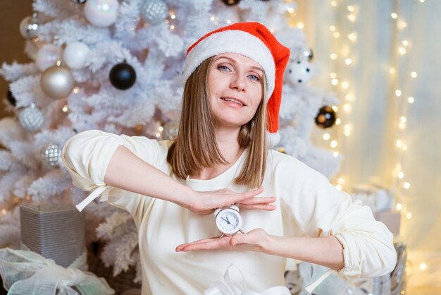 Felice bella giovane donna con un cappello rosso di Babbo Natale in un maglione bianco che punta a un orologio rotondo su...