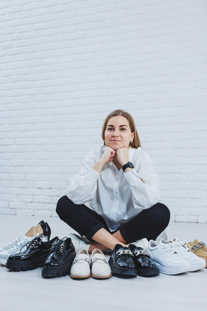 Felice bella giovane donna con un sorriso in un vestito alla moda con una camicia bianca e pantaloni neri si siede sul pavimento tra le scarpe e sceglie un nuovo paio di scarpe per se stessa