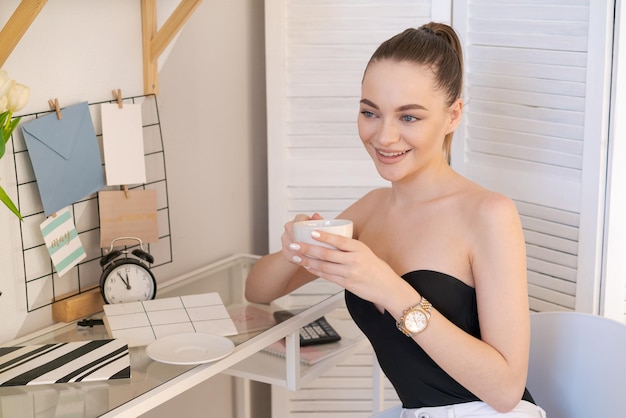 Felice bella giovane donna si siede alla sua scrivania a casa con una tazza di caffè in lei