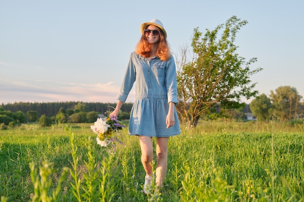 Felice bella giovane ragazza adolescente che cammina sul prato in abito cappello occhiali con bouquet di fiori