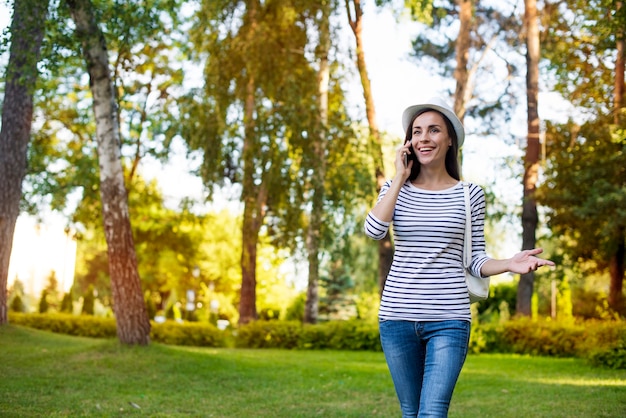 Felice bella giovane studentessa in un cappello con smart phone e zaino mentre cammina all'aperto nel parco