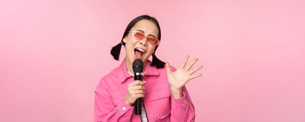 Felice bella ragazza asiatica che canta con il microfono utilizzando il microfono godendo il karaoke in posa su sfondo rosa studio