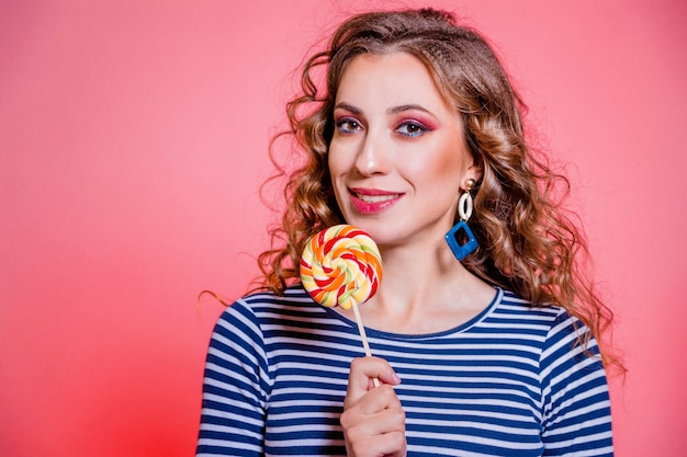 Felice bella ragazza bruna con trucco rosso, capelli ricci e un maglione a strisce blu in posa su uno sfondo rosso. Foto orizzontale