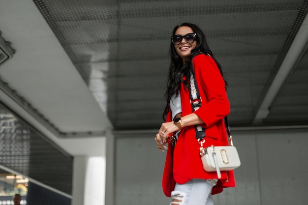 Felice bella ragazza con un sorriso che indossa occhiali da sole alla moda in un blazer rosso alla moda con una borsa alla moda sta camminando in città su uno sfondo urbano