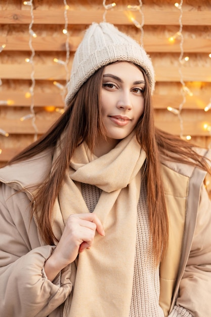 Felice bella ragazza in capispalla alla moda con un cappello lavorato a maglia, una giacca invernale e un'elegante sciarpa vicino a una parete di legno all'esterno