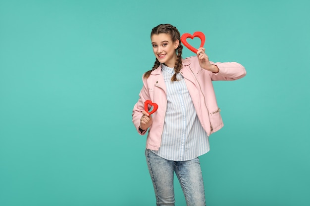 Felice bella ragazza in stile casual, capelli a codino e giacca rosa, in piedi e con in mano forme di cuore rosso e guardando la telecamera con un sorriso a trentadue denti, al coperto, isolato su sfondo blu o verde