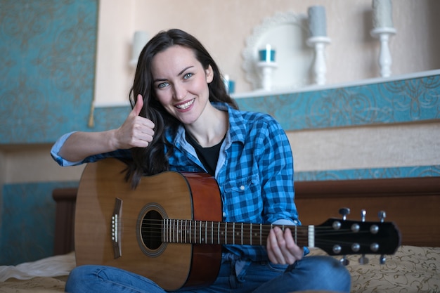 felice bruna allegra si siede sul letto con una chitarra acustica e mostra il pollice in mano