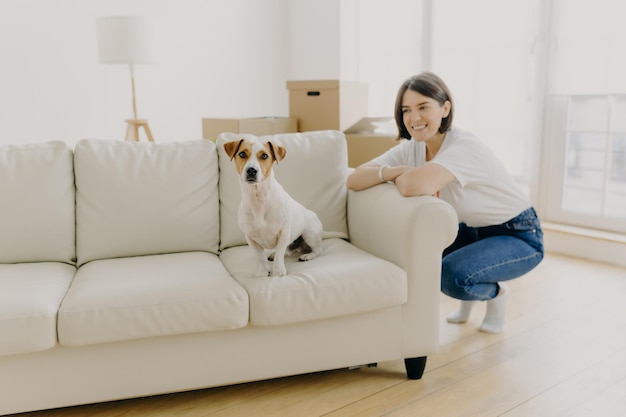 Felice bruna femmina in abbigliamento casual si appoggia sul lato del divano bianco pedigree cane domestico pone sul divano in appartamento vuoto vicino al proprietario della donna scatole disimballate in background Persone e concetto di casa