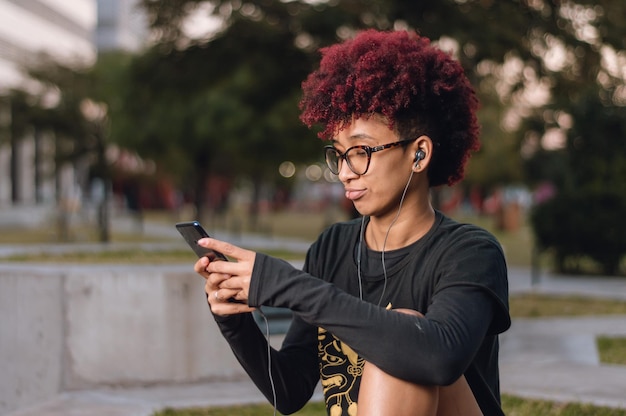 Felice bruna giovane donna latina con afro seduto all'aperto controllando il suo telefono