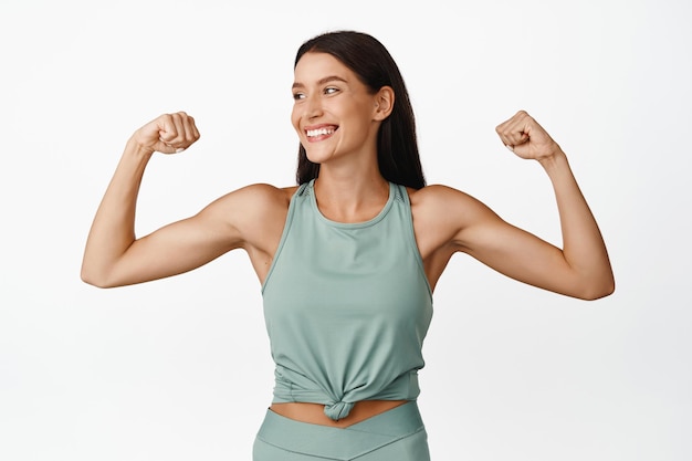 Felice bruna sportiva flettendo il bicipite sorridente e guardando da parte Fitness donna sentirsi sano e forte allenamento in palestra indossando activewear sfondo bianco