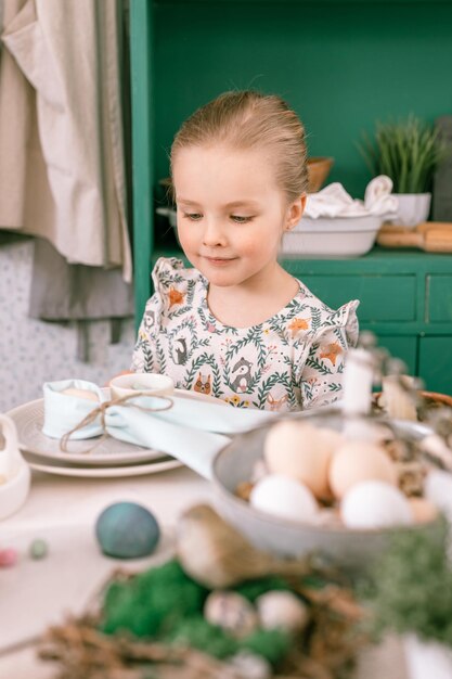 Felice candida bambina di quattro anni divertirsi pronto primavera Pasqua vacanze a casa cucina aiuta sua madre a decorare la tavola festiva con uova di Pasqua e prodotti da forno e caramelle per pranzo o cena