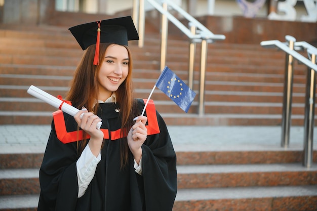 Felice carina brunetta caucasica ragazza laureata sorride Lei è in un mortaio nero con nappa rossa in abito con un bel diploma di capelli ricci castani in mano