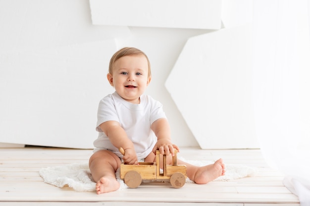 Felice carino piccolo bambino di sei mesi in una maglietta bianca e pannolini si siede su uno sfondo chiaro a casa e gioca con una macchina da scrivere in legno, spazio per il testo