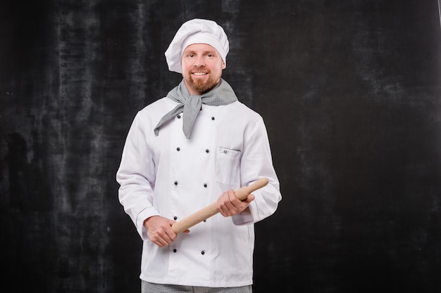 Felice chef barbuto in uniforme bianca che tiene il mattarello in legno mentre in piedi davanti alla telecamera su sfondo nero