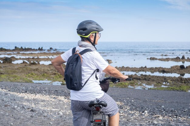 Felice ciclista donna anziana che indossa casco e zaino in piedi lungo la spiaggia del mare guardando l'orizzonte sull'acqua