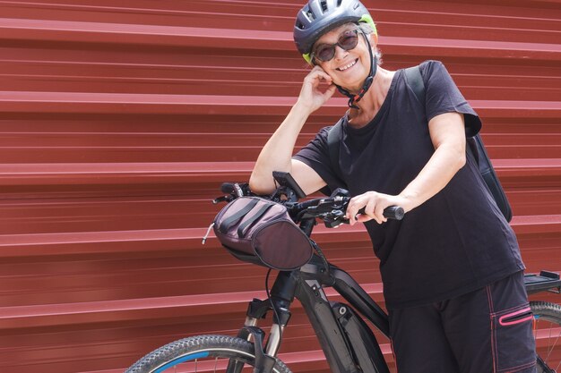 Felice ciclista donna anziana in un'escursione all'aperto vicino al pannello di metallo rosso che si gode uno stile di vita sano