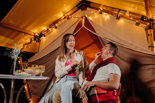 Felice coppia adorabile che si rilassa nel glamping la sera d'estate e mangia uva vicino al falò accogliente Tenda da campeggio di lusso per attività ricreative all'aperto e attività ricreative Concetto di stile di vita