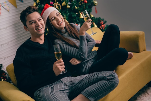 Felice coppia asiatica brindando champagne insieme seduti sul divano la sera con un albero di Natale e luci sullo sfondo Fidanzato e fidanzata alla notte della vigilia di Natale