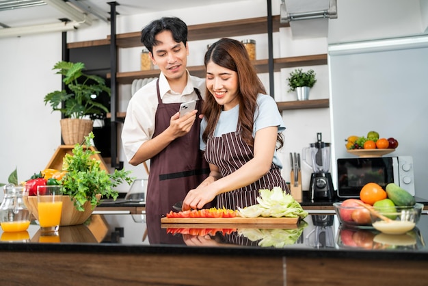 Felice coppia asiatica che indossa grembiuli L'uomo vive dal telefono cellulare durante la donna prepara un'insalata vegetariana sana con verdure fresche come carote, pomodori, cavoli e quercia verde nella cucina di casa