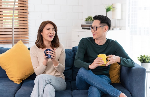 Felice coppia asiatica uomo e donna sta trascorrendo il fine settimana insieme sul divano in casa, rilassandosi e godendosi il caffè.