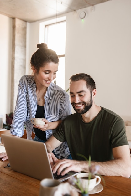 felice coppia bruna uomo e donna che bevono caffè e lavorano insieme al computer portatile mentre era seduto al tavolo a casa