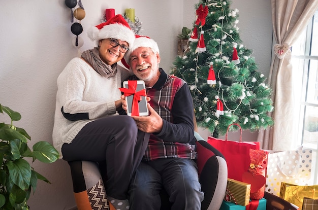 Felice coppia di anziani con il berretto di Babbo Natale seduti insieme nella stessa poltrona. Con in mano un tablet ricevuto come regalo di Natale. Regali e albero di Natale in sottofondo