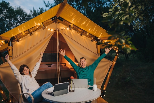 Felice coppia di famiglia freelance che lavorano laptop su un'accogliente tenda glamping in serata estiva Tenda da campeggio di lusso per vacanze e vacanze all'aperto Concetto di stile di vita