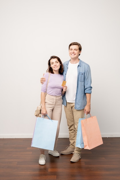 Felice coppia di giovani acquirenti con paperbags e carta di credito in piedi dal muro in isolamento in studio