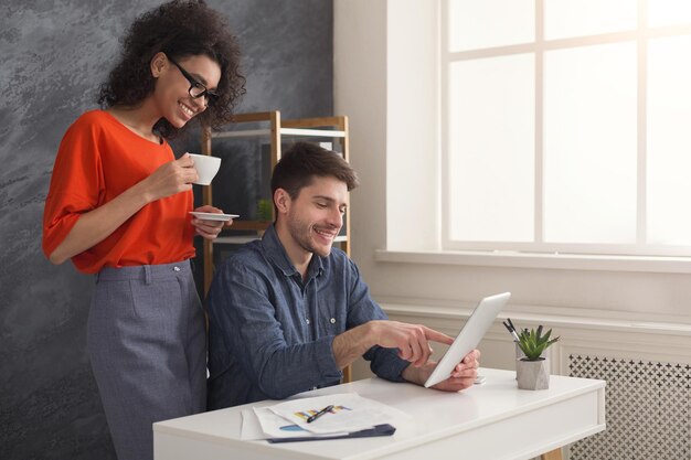 Felice coppia di giovani partner commerciali che lavorano in un ufficio moderno. Due colleghi sorridenti che discutono di un nuovo progetto mentre lavorano su tablet digitale, donna che beve caffè, spazio copia