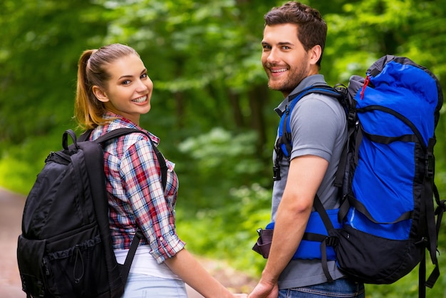 Felice coppia di turisti. Bella giovane coppia di innamorati che trasportano zaini e si guardano le spalle con un sorriso mentre camminano lungo il sentiero della foresta