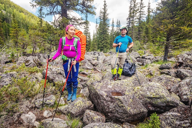 Felice coppia di viaggiatori con bastoncini da trekking e un grande zaino cammina lungo un sentiero tra enormi pietre e massi in un parco nazionale con un fiume di montagna sullo sfondo