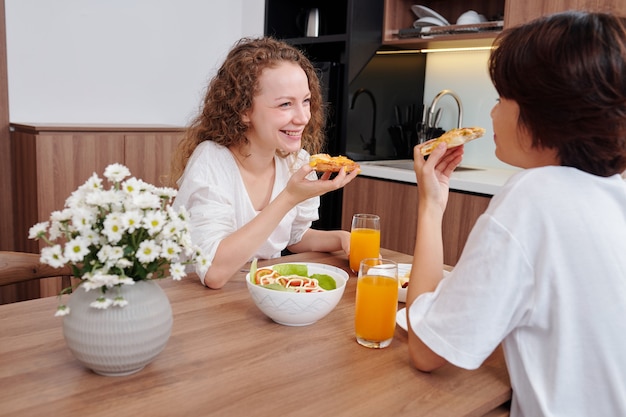 Felice coppia lesbica che si gode una gustosa pizza con insalata e succo d'arancia per cena, ridendo e discutendo di notizie