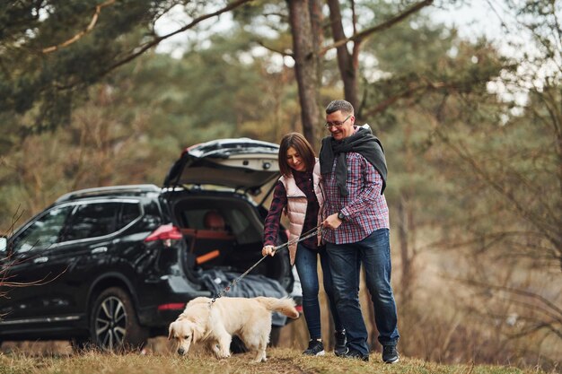 Felice coppia matura fare una passeggiata con il loro cane nella foresta autunnale o primaverile vicino a un'auto moderna