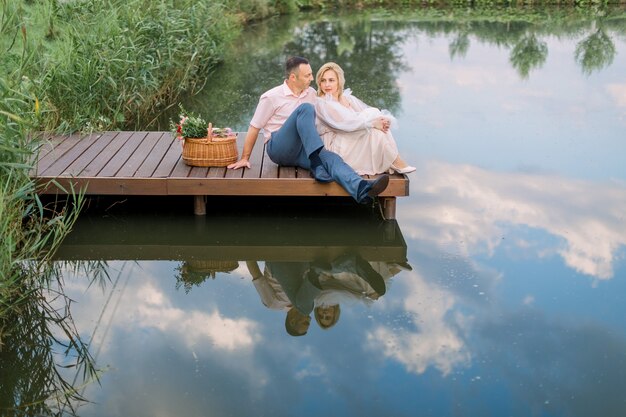 Felice coppia matura romantica seduta su un ponte di legno vicino al lago o allo stagno all'aperto, abbracciando e godendosi il loro appuntamento. Celebrazione dell'anniversario di matrimonio