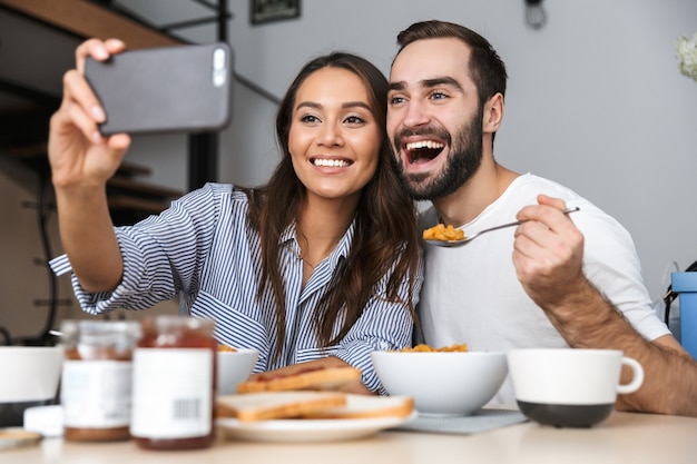 Felice coppia multietnica facendo colazione in cucina, prendendo un selfie
