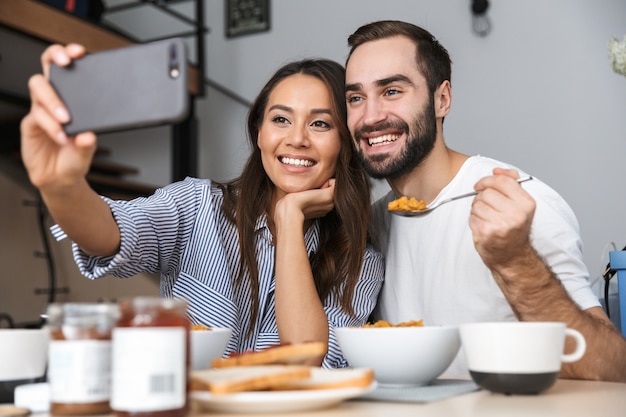 Felice coppia multietnica facendo colazione in cucina, prendendo un selfie