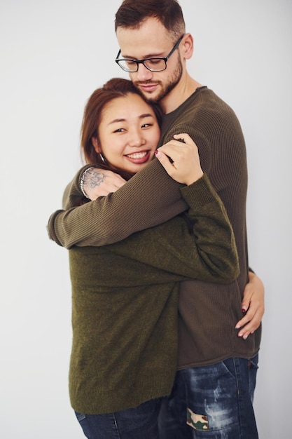 Felice coppia multietnica in abiti casual che si abbracciano al chiuso in studio. Ragazzo caucasico con la ragazza asiatica.